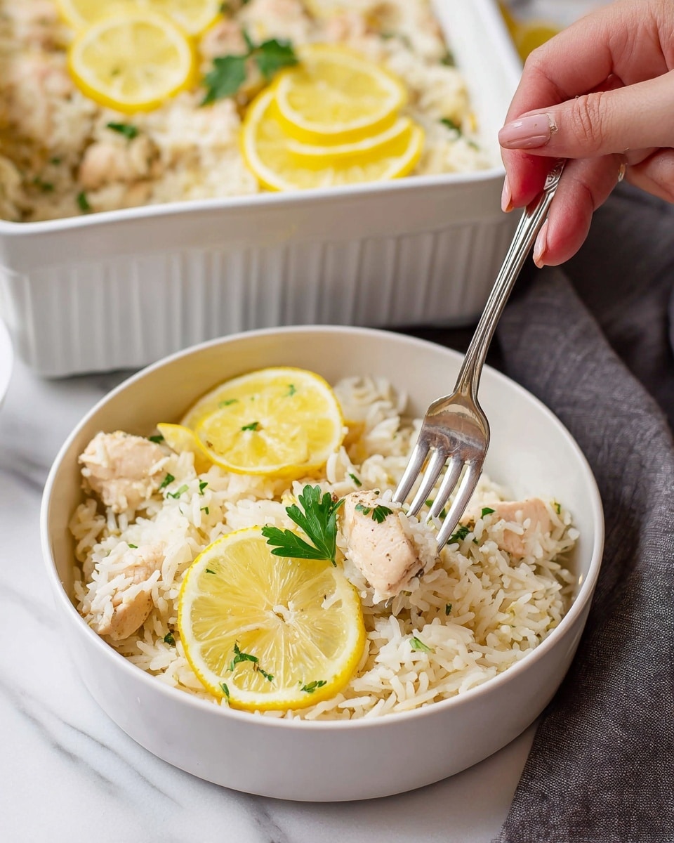 A white bowl filled with a mix of white rice and small pieces of cooked chicken layered evenly, topped with bright yellow lemon slices on one side and a small green parsley leaf near the center, with a fork pressing down on a chicken piece; behind it is a white ceramic baking dish with more of the same rice and chicken mixture garnished with lemon slices and a parsley sprig, set on a white marbled surface, with a gray cloth nearby and a woman's hand holding the fork. photo taken with an iphone --ar 4:5 --v 7