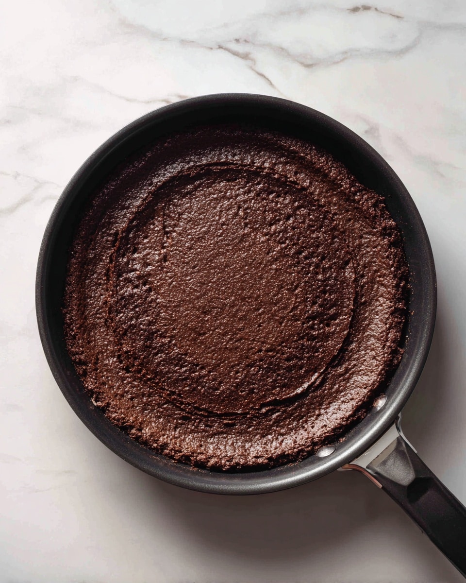A single dark brown flat layer of thick pancake batter cooked unevenly in a black pan, showing rough texture with some crinkled and folded edges, and slight cracking on the surface. The pan handle is partially visible on the top right side, and the background consists of a white marbled surface. photo taken with an iphone --ar 4:5 --v 7