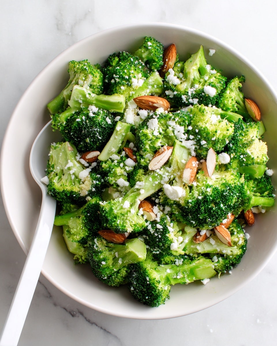 The image shows a white bowl filled with fresh green broccoli florets on a white marbled surface. The broccoli pieces are vibrant and bright green with small bits of white cheese sprinkled over them, adding texture and contrast. Scattered among the broccoli are whole and sliced almonds, providing a light brown color and crunchy texture to the dish. A white spoon is placed inside the bowl on the left side, ready for serving. The overall look is fresh, healthy, and simple. Photo taken with an iphone --ar 4:5 --v 7