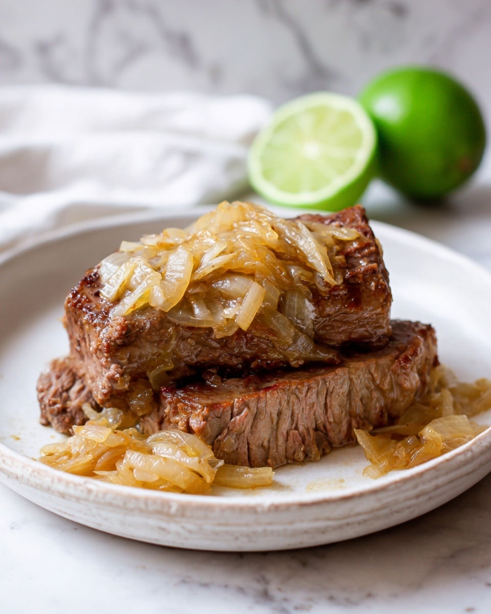 The image shows a white plate with two pieces of cooked brown beef stacked on each other. On top of the beef, there is a generous layer of light golden cooked onions with soft texture, some falling around the meat on the plate. In the background, there is a half lime placed on a white marbled surface. The photo taken with an iphone --ar 4:5 --v 7