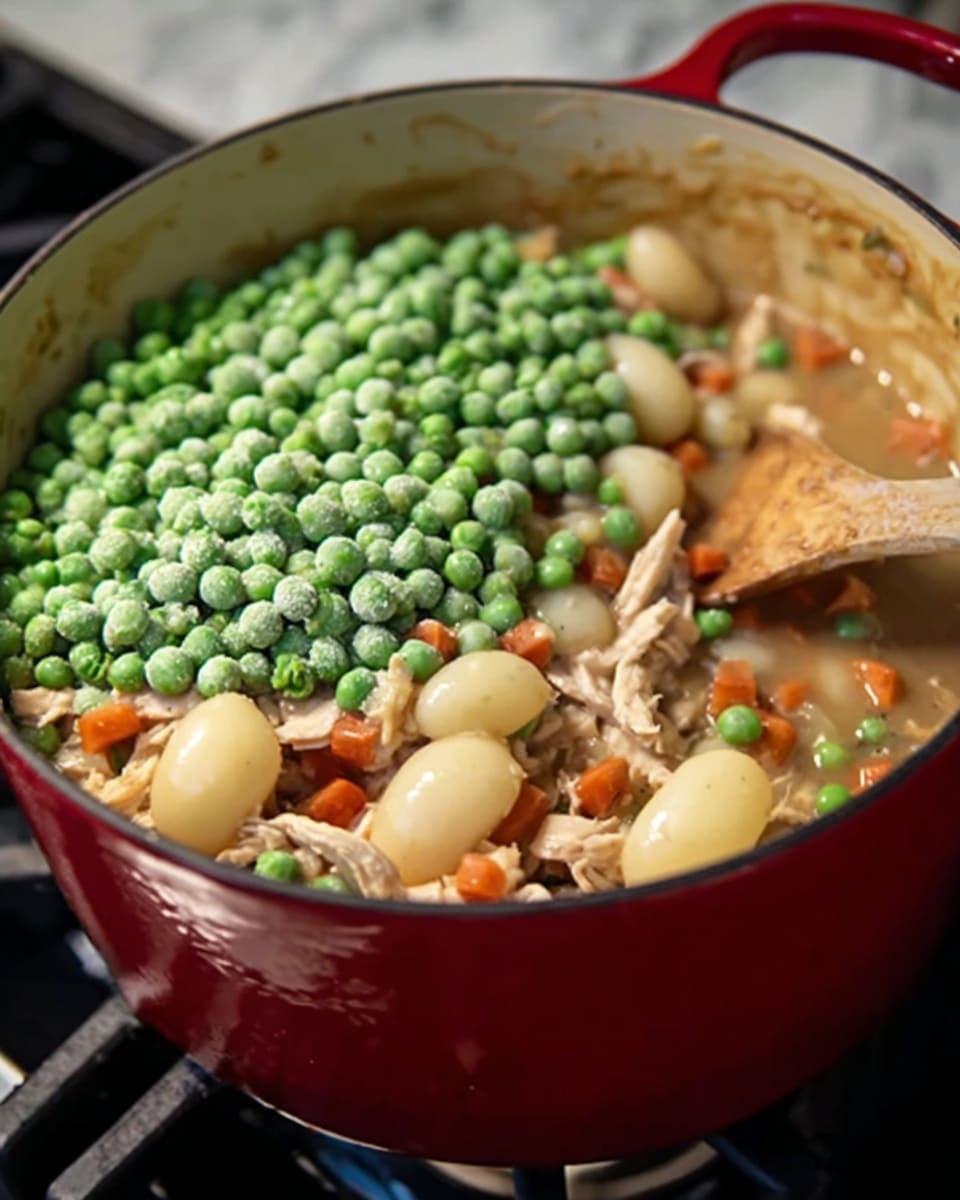 A close-up image shows a thick stew in a red pot on a white marbled stove. The stew has several layers: the bottom layer is light brown sauce with visible small orange carrot pieces and shredded light beige chicken mixed with soft, pale round potato pieces. On top of this, a generous heap of bright green frozen peas adds contrast and texture. A wooden spoon is partly dipped in the stew, suggesting it is being stirred. The background is softly blurred, focusing closely on the pot and stew. photo taken with an iphone --ar 4:5 --v 7