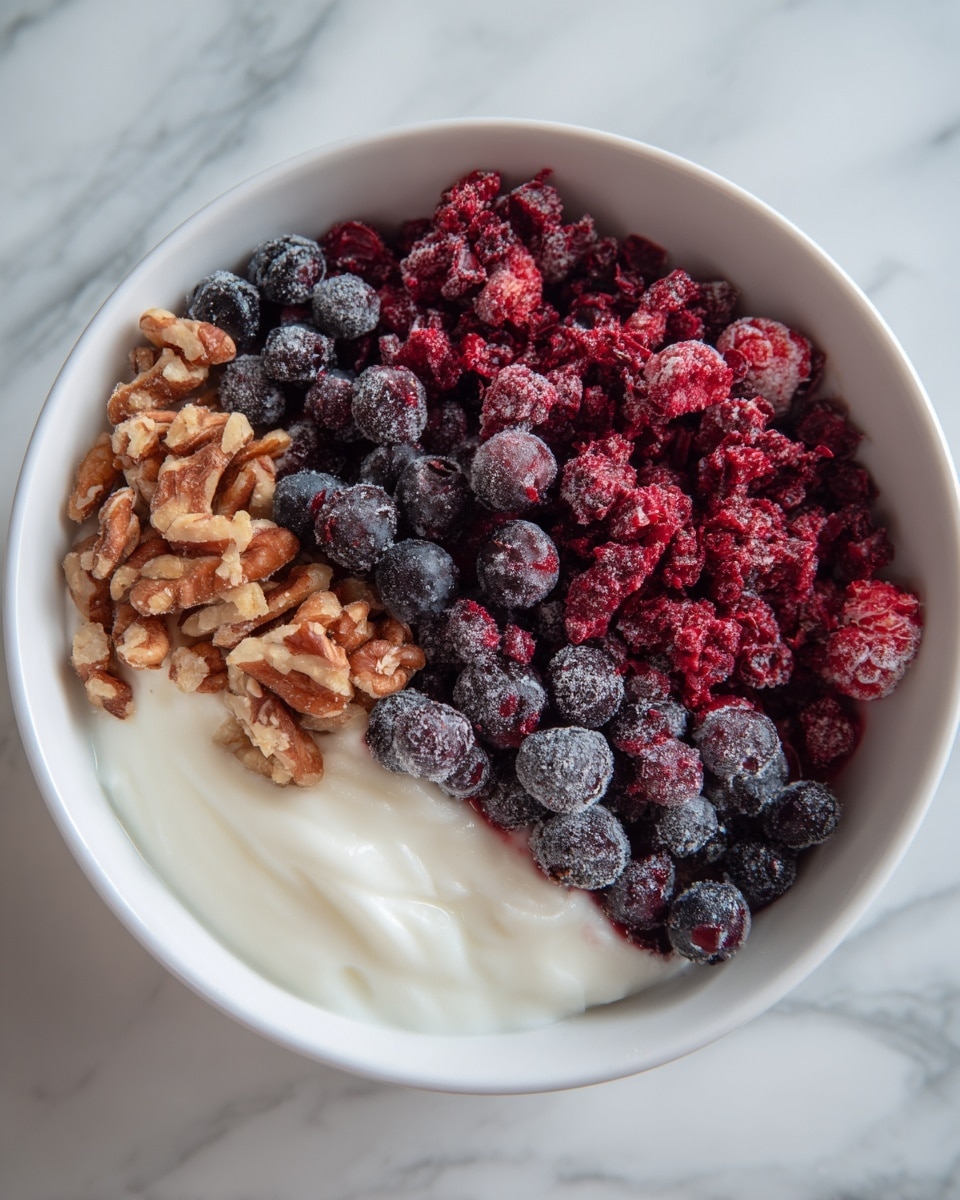 The image shows a white bowl filled with three visible layers. The bottom layer is white, creamy yogurt with a smooth texture. On top of the yogurt is a layer of brown, chopped nuts spread mainly on the left side. The top layer consists of a large pile of frozen mixed berries in various shades of red, deep purple, and dark blue, covering most of the right side and center of the bowl. The berries look frosty and have a rough texture. The bowl is placed on a white marbled surface. photo taken with an iphone --ar 4:5 --v 7