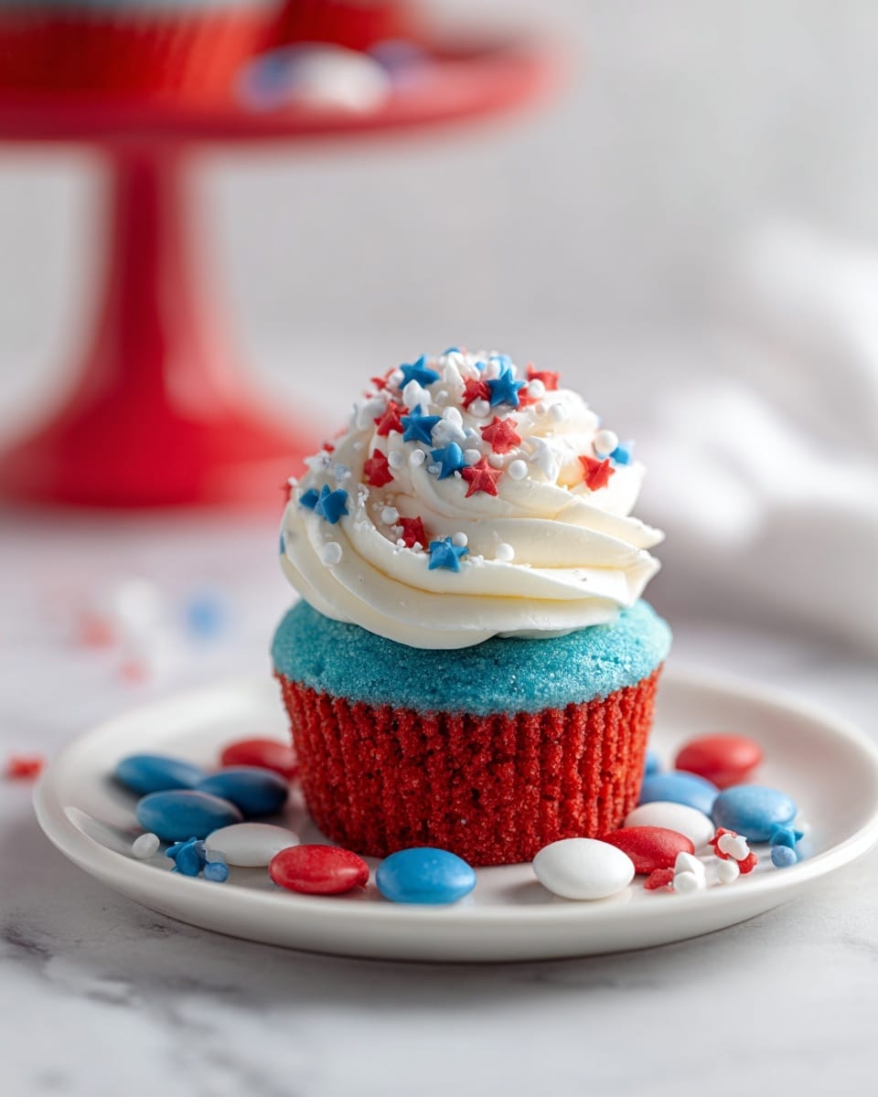 A small cupcake with three colorful layers sits on a white plate over a white marbled surface. The bottom layer is bright red with a slightly rough texture, the middle layer is creamy white and smooth, and the top layer is bright blue and soft looking. On top, there is a swirl of white whipped cream decorated with red, blue, and white round and star-shaped sprinkles. Around the cupcake on the plate are matching red, blue, and white small round candy pieces. In the blurry background, a red cake stand is visible. Photo taken with an iphone --ar 4:5 --v 7