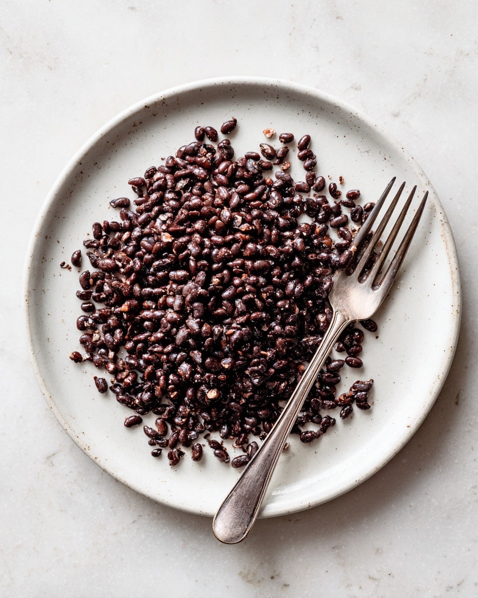 A white round plate filled with a single layer of small, dark brown to almost black beans that are slightly broken and crushed, showing some lighter brown bits mixed in. There is a vintage silver fork placed on the right edge of the plate with a slight worn texture. The plate is placed on a white marbled surface that adds soft contrast to the food and gives a clean, natural look. Photo taken with an iphone --ar 4:5 --v 7
