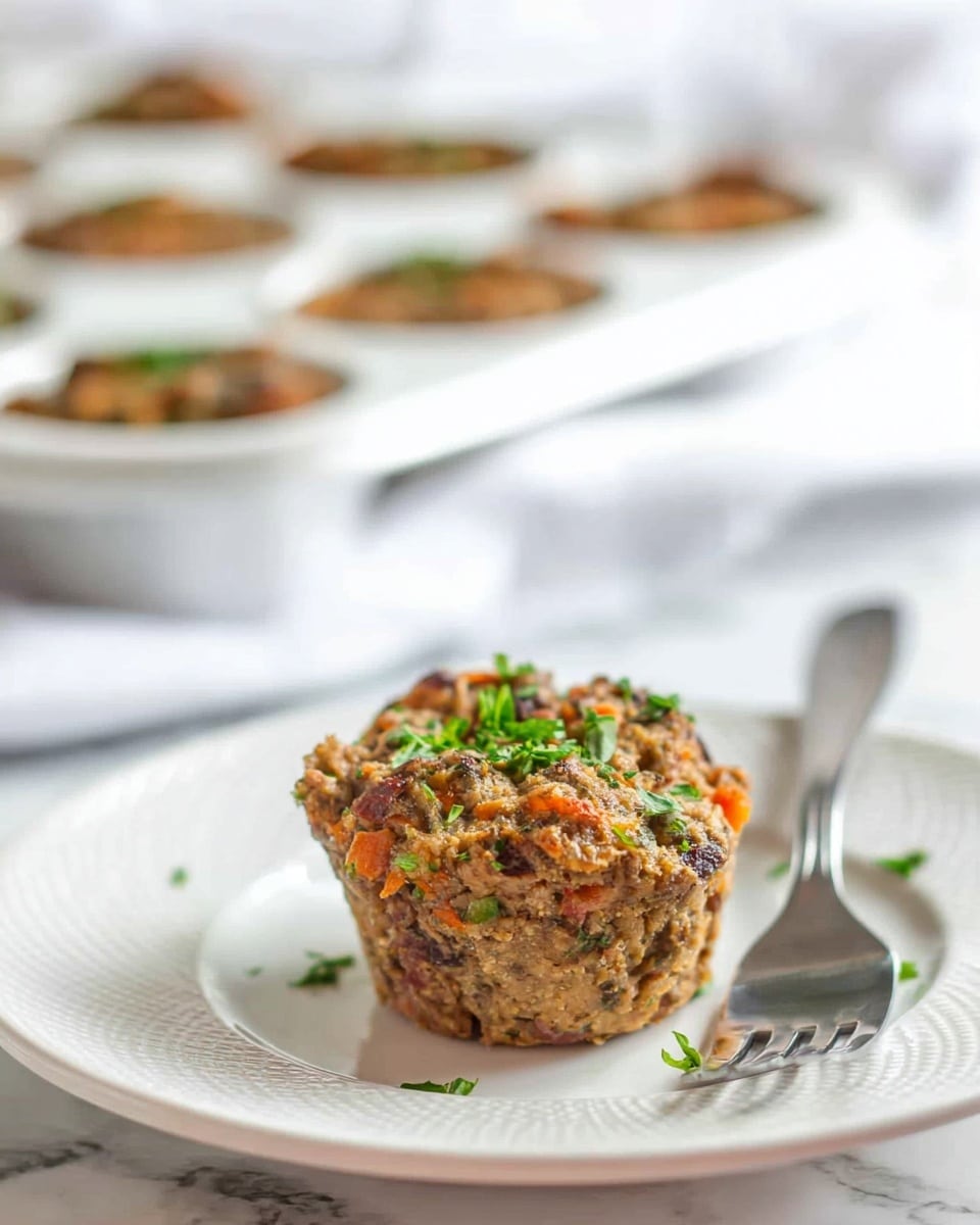 The image shows one small muffin-shaped meat dish placed on a white plate with a subtle textured pattern. The meat muffin has a rough, dense texture with bits of orange and green visible inside, topped with small chopped green herbs. Behind the plate, there is a blurred white muffin tin holding more of the same meat muffins. The surface beneath everything is a white marbled texture. A silver fork rests beside the muffin on the plate. photo taken with an iphone --ar 4:5 --v 7