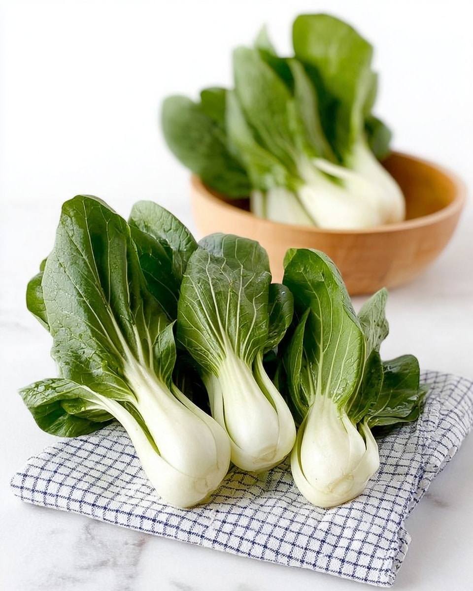 The image shows fresh baby bok choy placed on a white and blue checkered cloth in the front, with three bunches closely arranged. The bok choy has bright white stems that are smooth and thick, leading up to dark green, slightly wrinkled leaves that look crisp and fresh. In the background, another bunch of bok choy is shown inside a light-colored wooden bowl. The scene is set against a clean white marbled surface. photo taken with an iphone --ar 4:5 --v 7