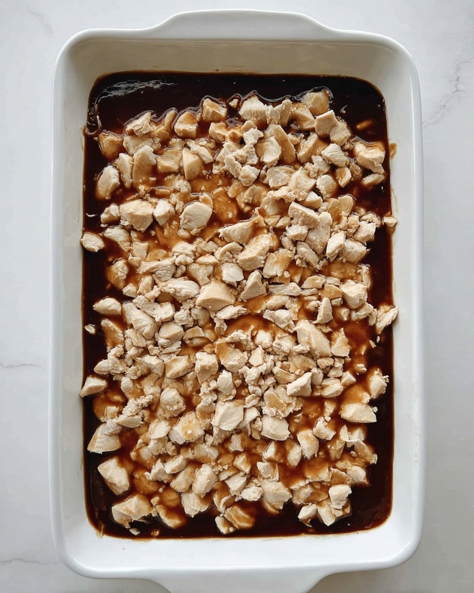 A white rectangular baking dish filled with a two-layer dish; the bottom layer is dark brown sauce with a smooth texture that covers the base fully, topped with an even layer of small, pale beige pieces of cooked chicken scattered uniformly across the surface. The dish is set on a white marbled background. Photo taken with an iphone --ar 4:5 --v 7