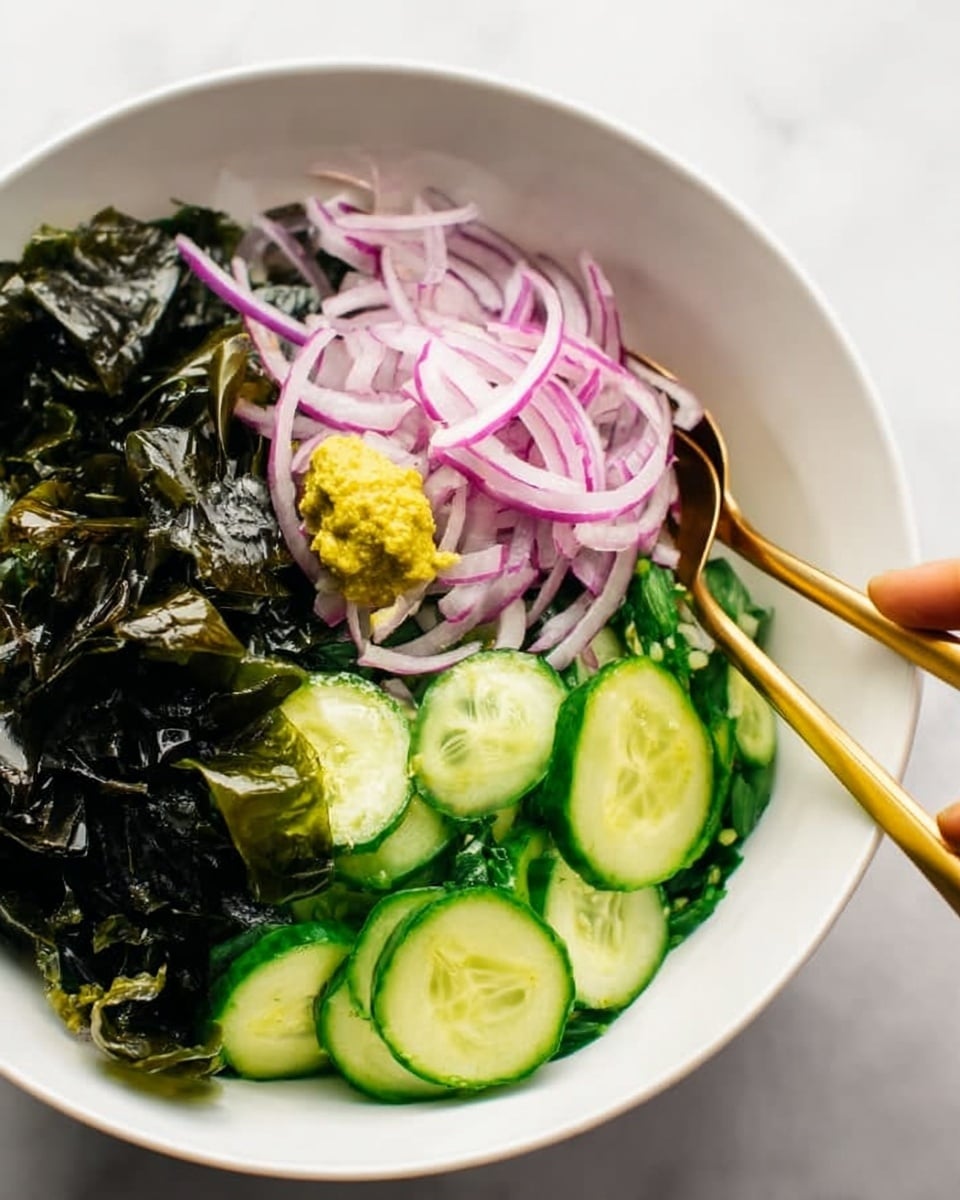 A white bowl filled with a fresh salad showing three main layers: bright green cucumber slices at the bottom with a bumpy texture, thin slices of purple onion placed in the middle, and dark green seaweed sheets on the side with a shiny texture. On top of the onions, there is a small dollop of yellow paste. Two golden spoons stand in the bowl from opposite sides, one of them touched by a woman's hand. The bowl sits on a white marbled surface. photo taken with an iphone --ar 4:5 --v 7