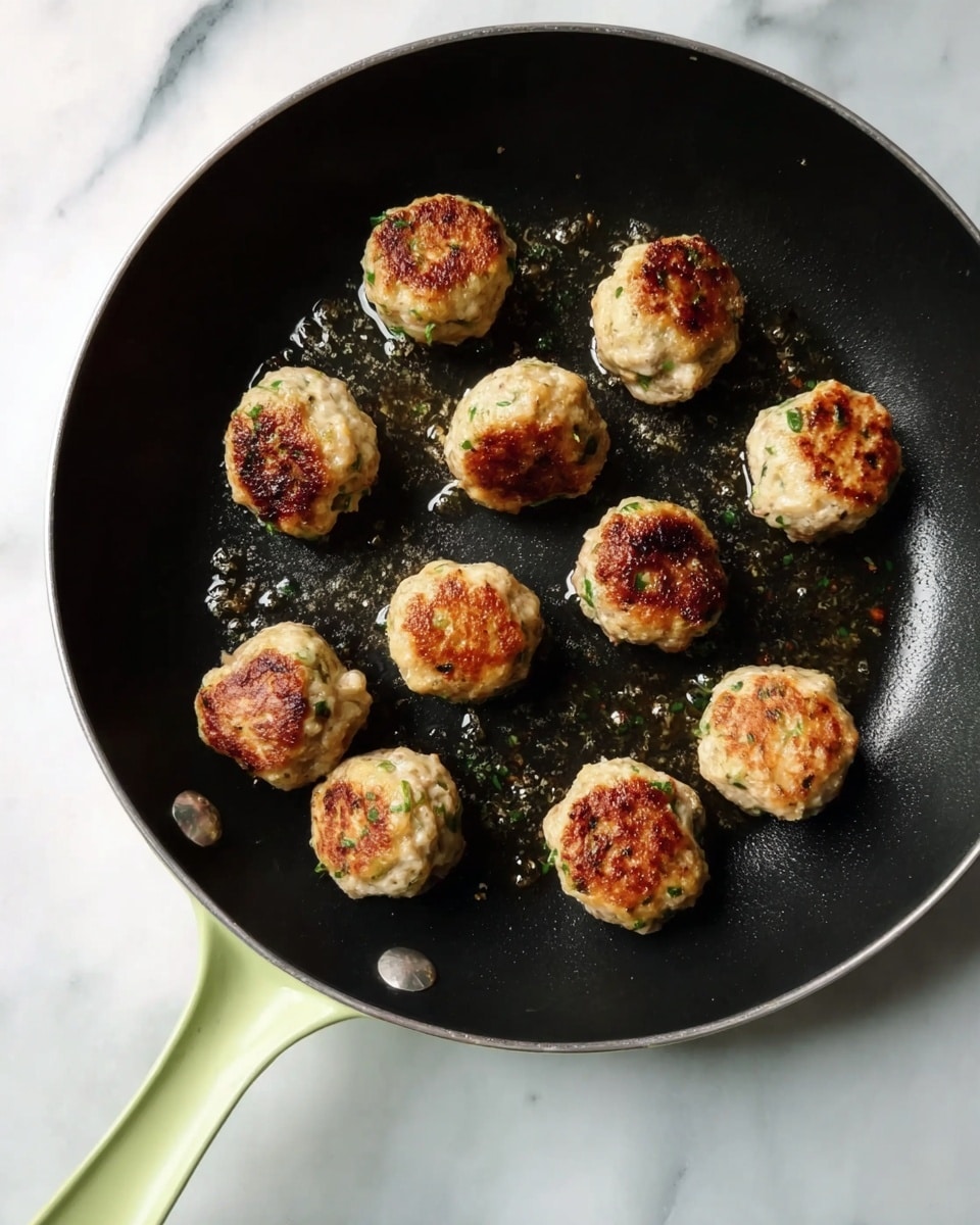 A black pan with a light green handle holds eleven small, round browned meatballs, each showing a golden-brown crust with visible bits of green herbs throughout. The meatballs are scattered mostly toward the left and right sides of the pan, which has slight oil glistening around them. The pan rests on a white marbled surface with soft gray veins. Photo taken with an iphone --ar 4:5 --v 7