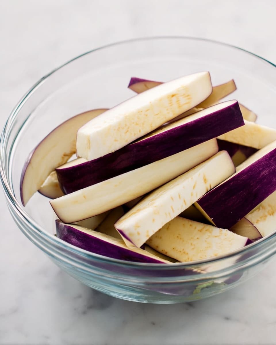 A clear glass bowl holds several long, thick slices of eggplant, mostly white with deep purple skin visible on some edges. The eggplant pieces are stacked unevenly in the bowl, showing smooth and slightly spotted pale flesh. This bowl is set on a white marbled surface, with soft natural light highlighting the textures and colors clearly. Photo taken with an iphone --ar 4:5 --v 7