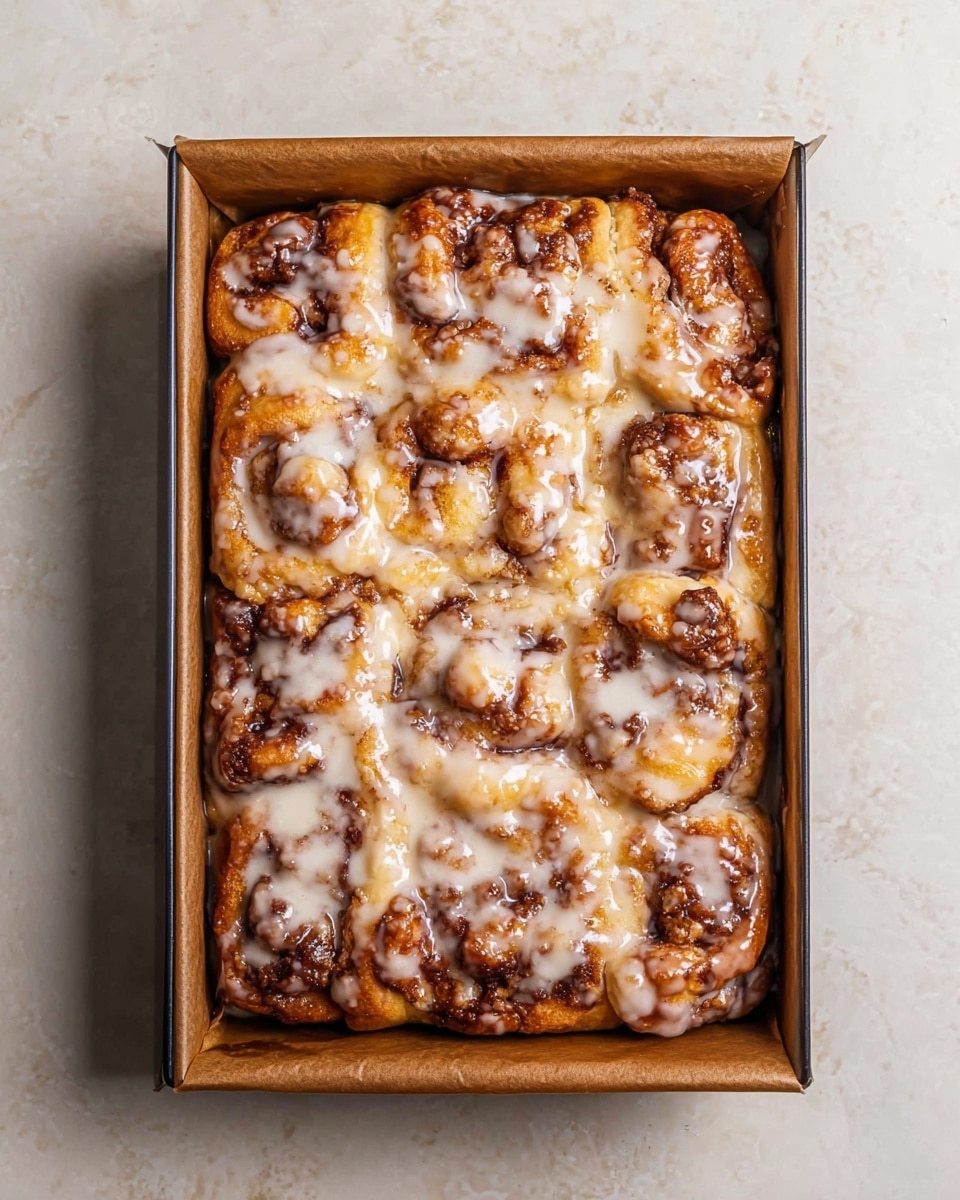 A rectangular baked cinnamon roll tray is shown from above, filled with soft, golden-brown rolls that have a textured, slightly crispy top with darker cinnamon spots. The rolls are thick and uneven, closely packed to fill the tray. They are covered with a layer of white, shiny icing that drips slightly between the rolls, giving a sticky, sweet look. The tray has brown parchment paper lining the edges, and the entire scene is set on a white marbled surface. photo taken with an iphone --ar 4:5 --v 7