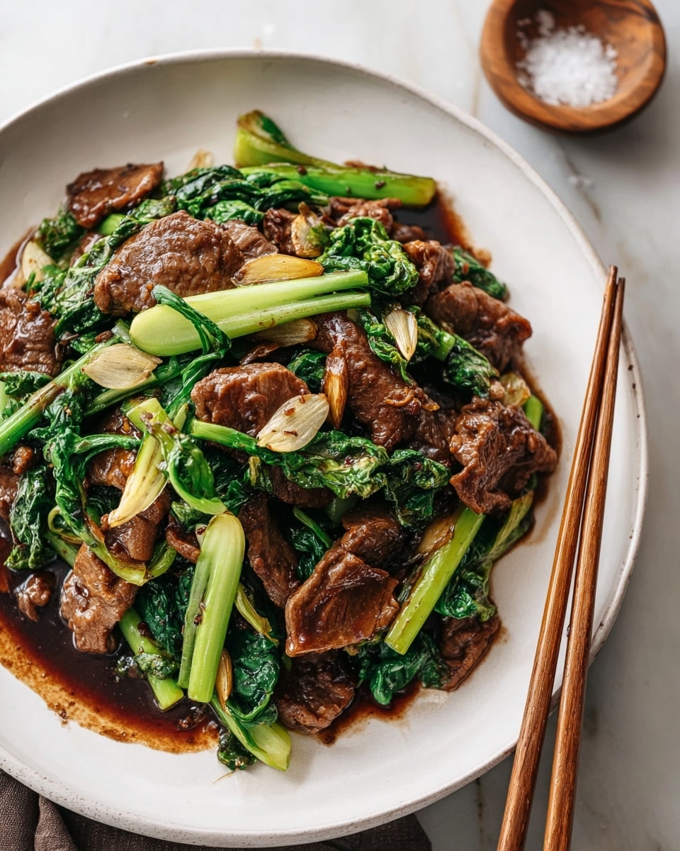 A white plate holds a dish with about three layers visible: the bottom layer is a dark brown sauce spread unevenly on the plate; above it are slices of brown cooked meat with a shiny texture, irregular in shape and size; mixed throughout are bright green leafy vegetables and chunks of green stems that add freshness and color contrast; scattered on top are thin slices of light golden garlic. A pair of brown wooden chopsticks rests on the side of the plate, with a soft white marbled surface underneath. In the top right corner, a small wooden bowl with coarse salt is partially visible. Photo taken with an iphone --ar 4:5 --v 7