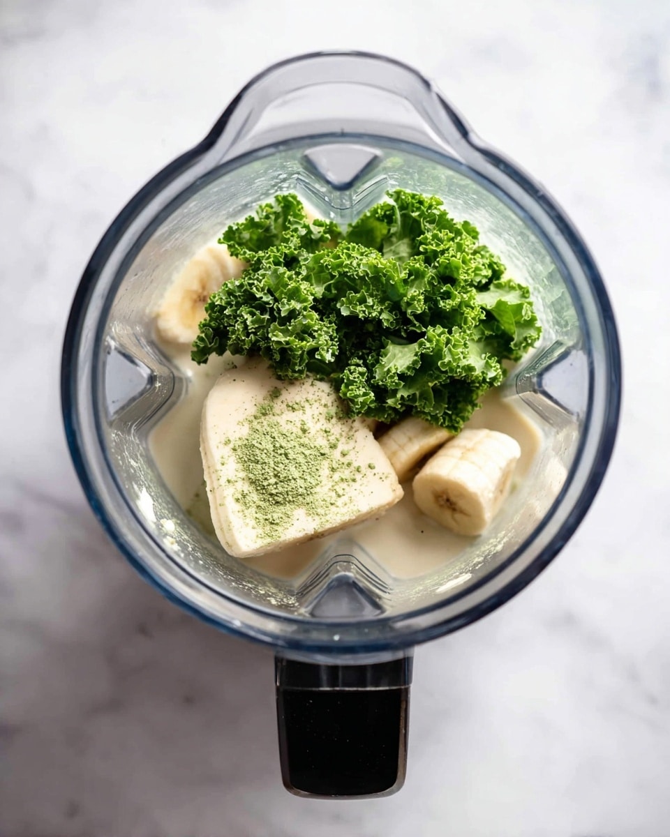 A clear blender jar with a black handle is shown from above, filled with three layers of ingredients: the bottom layer is a light creamy beige liquid, the middle layer has two pieces of pale, peeled banana, and the top layer is bright green leafy kale with some fine green powder sprinkled on it. The blender jar sits on a white marbled surface. photo taken with an iphone --ar 4:5 --v 7