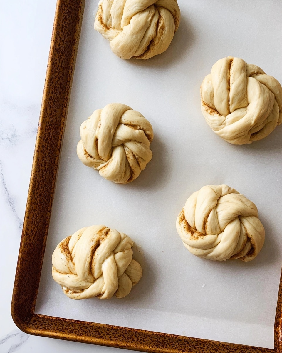 The image shows five twisted dough buns on a baking tray lined with white parchment paper. Each bun is shaped like a knotted flower, with two or three layers of dough twisted together, showing shades of light beige and hints of cinnamon brown spiral inside the folds. The dough looks soft but not yet baked, with smooth and slightly shiny surfaces. The baking tray has a textured edge and is set on a white marbled surface. Photo taken with an iphone --ar 4:5 --v 7