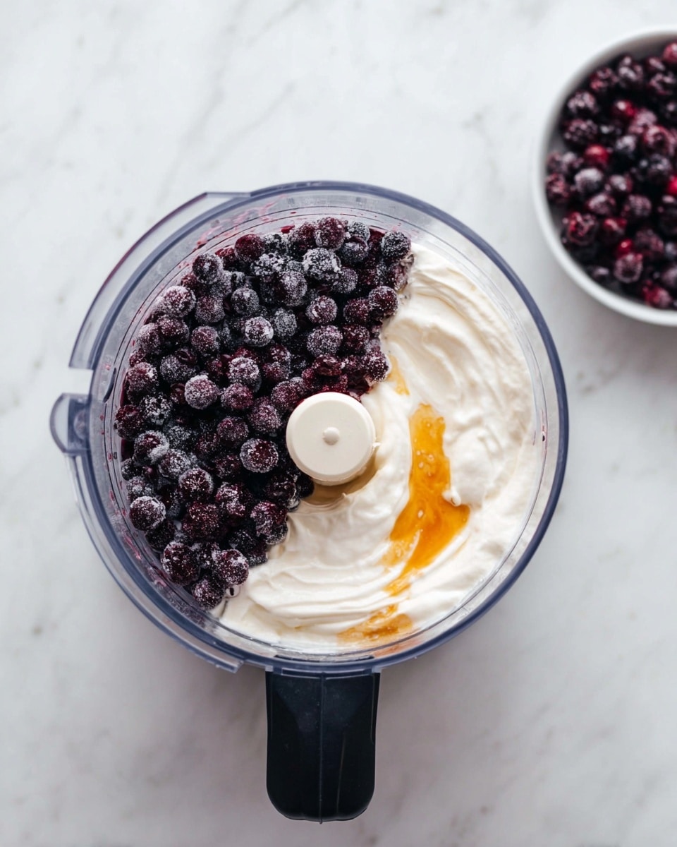 The image shows a clear food processor bowl with two main layers inside. One half of the bowl is filled with small, dark purple frozen berries with a slightly frosty texture. The other half holds a thick, creamy white layer of yogurt or cream with a small amount of golden honey or syrup on top, blending lightly into the creamy layer. The background is a white marbled surface with a small white bowl filled with more frozen berries placed nearby. photo taken with an iphone --ar 4:5 --v 7
