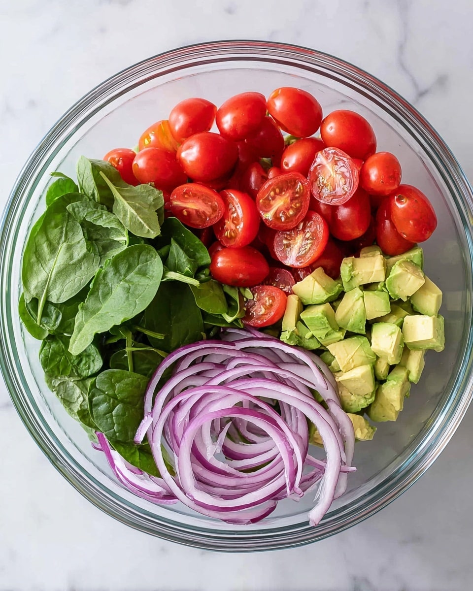 A clear glass bowl sits on a white marbled surface holding four main layers of fresh ingredients separated by color and texture. The top left layer is made of bright red cherry tomatoes, some whole and some cut in half, showing their juicy inside. Below the tomatoes, at the bottom left, is a bed of fresh green spinach leaves with a soft, slightly shiny texture. The bottom right layer features thin, curved slices of purple onion with white edges showing their crisp layers. The top right layer is a mix of diced light yellow avocado pieces with a creamy texture. photo taken with an iphone --ar 4:5 --v 7