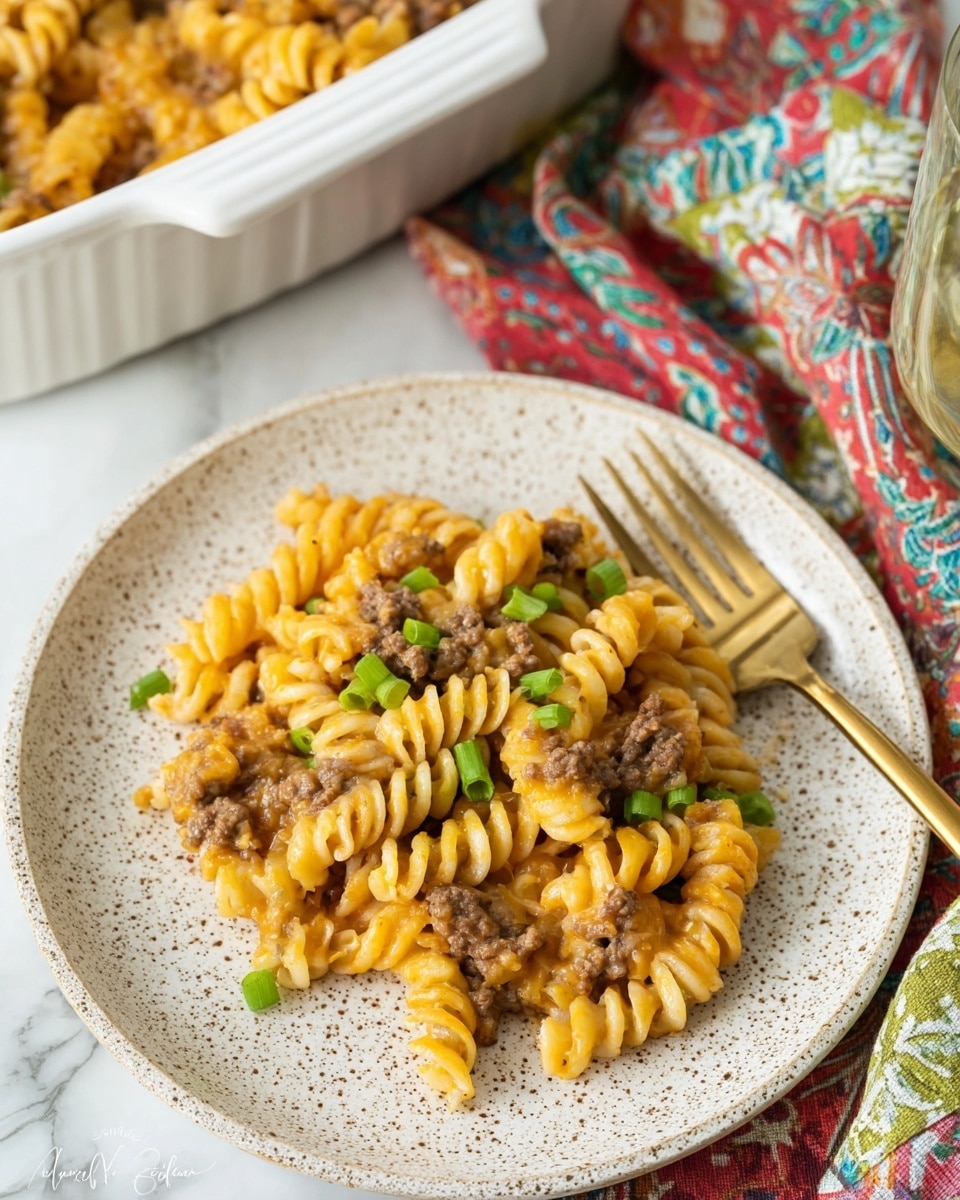 A close-up view of a white speckled plate filled with three layers of food: the first layer is light orange spiral pasta, the second layer is small chunks of brown cooked meat mixed throughout, and the third layer is chopped green onions scattered on top. The plate is on a white marbled surface with a gold-toned fork placed near the top edge of the plate. In the background, part of a white casserole dish filled with the same pasta mixture is visible along with a colorful cloth with red, green, and blue patterns. Photo taken with an iphone --ar 4:5 --v 7