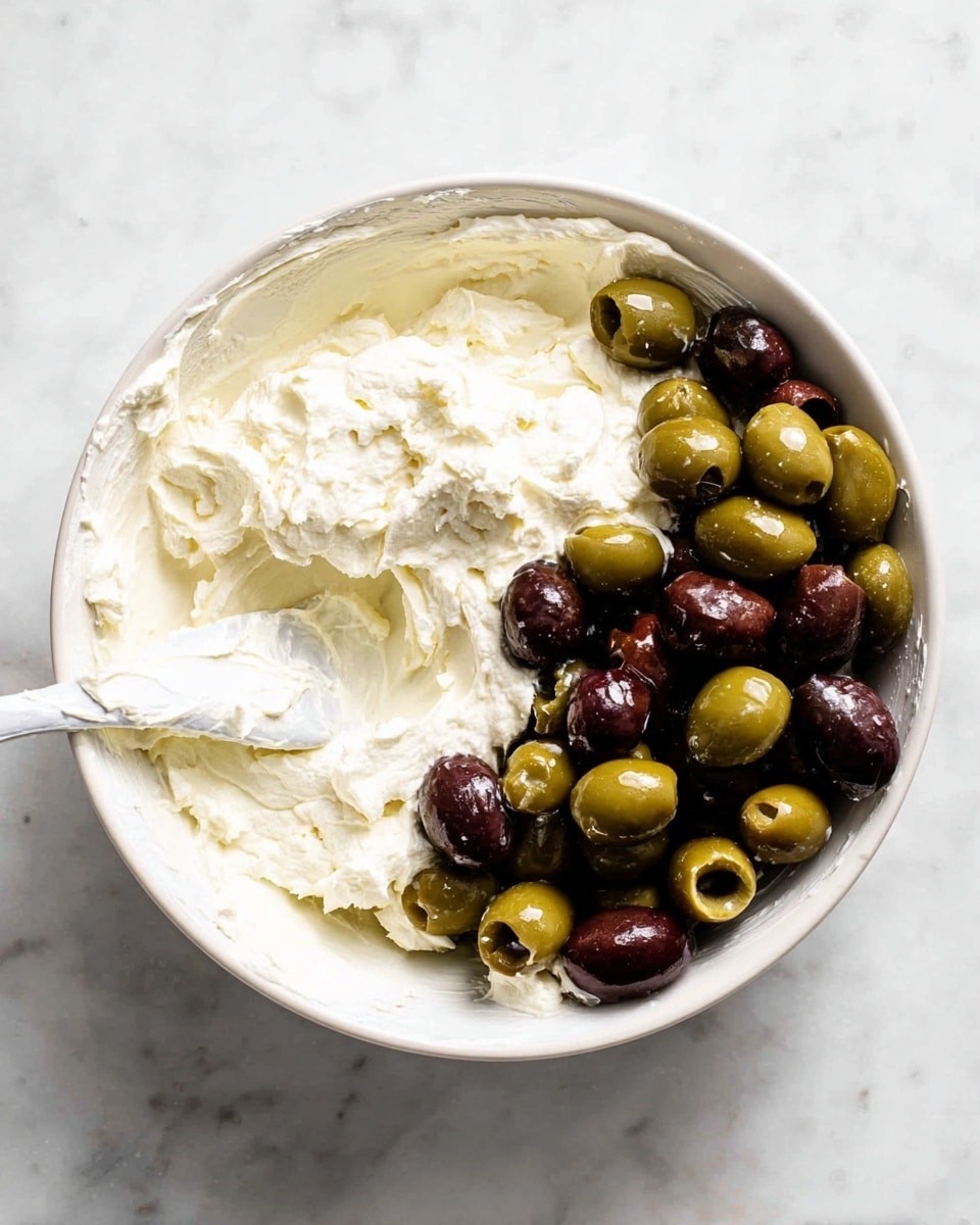A white bowl sits on a white marbled surface filled halfway with a thick, creamy white mixture on the left side, showing a soft and textured look. On the right side of the bowl, there is a pile of sliced green and dark purple olives, their shiny surfaces adding color contrast to the creamy mixture. A white spatula rests inside the bowl, partially covered in the creamy mixture, angled towards the left side. The image is bright and clear, with natural light highlighting the textures and colors photo taken with an iphone --ar 4:5 --v 7