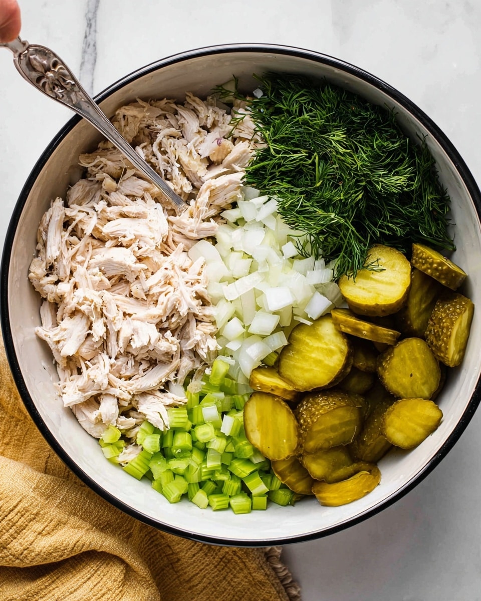 A large white bowl with a black rim holds layers of ingredients arranged in sections. At the bottom left, there is a layer of light beige shredded chicken with a soft texture. Next to it, on the bottom right, are small chopped light green celery pieces. Moving up to the top right section, there is a pile of sliced pickles with a shiny green and yellow look. At the top middle, lighter shredded chicken pieces sit beneath the pickles. To the left of the chicken, finely chopped white onions cover the area. Finally, the top left section features a bunch of fresh, deep green chopped dill herbs. A silver spoon and another utensil rest inside the bowl, and a woman's hand can just be seen holding one handle. The scene is set on a white marbled surface with a folded yellow cloth nearby. Photo taken with an iphone --ar 4:5 --v 7