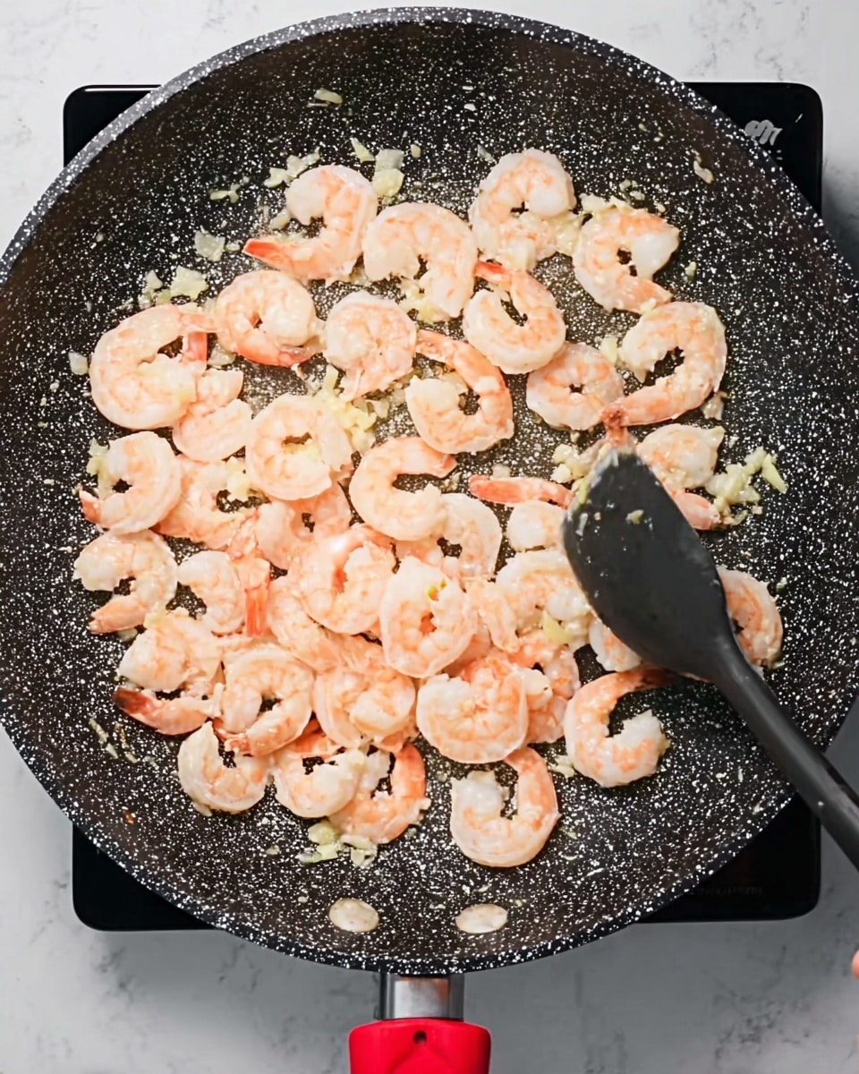 A close-up top view of a black speckled frying pan with a red handle filled with many shrimp cooking in oil. The shrimp are light pink and pale orange in color, spread evenly across the pan. Small bits of white garlic pieces are scattered among the shrimp. A woman's hand is holding a black spatula, visible on the right side of the pan stirring the shrimp. The background surface is a white marbled texture. Photo taken with an iphone --ar 4:5 --v 7