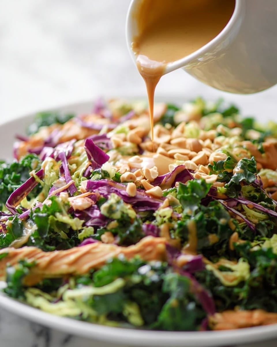 A close-up view shows a colorful salad on a white plate, with layers of green kale and spinach leaves, pieces of light purple cabbage, and light brown chicken strips mixed with shredded cheese and small peanut bits scattered on top. Above the salad, a white ceramic pitcher is pouring a smooth, creamy tan dressing onto the vegetables. The background is a white marbled texture. photo taken with an iphone --ar 4:5 --v 7