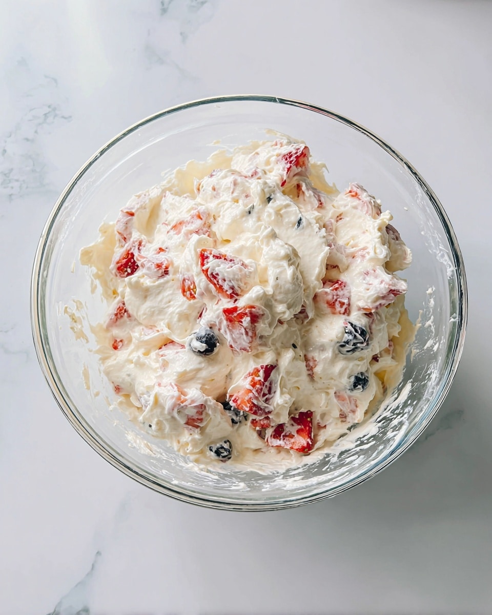 A clear glass bowl filled with a creamy white mixture, layered with fresh red strawberry pieces and whole dark blue blueberries evenly distributed throughout. The creamy texture looks soft and light, with small white marshmallows scattered inside. Around the bowl on a white marbled surface, there are whole strawberries with green leaves and loose blueberries, as well as mini white marshmallows. A bright blue textured cloth with white beads draped nearby adds color contrast. The photo taken with an iphone --ar 4:5 --v 7