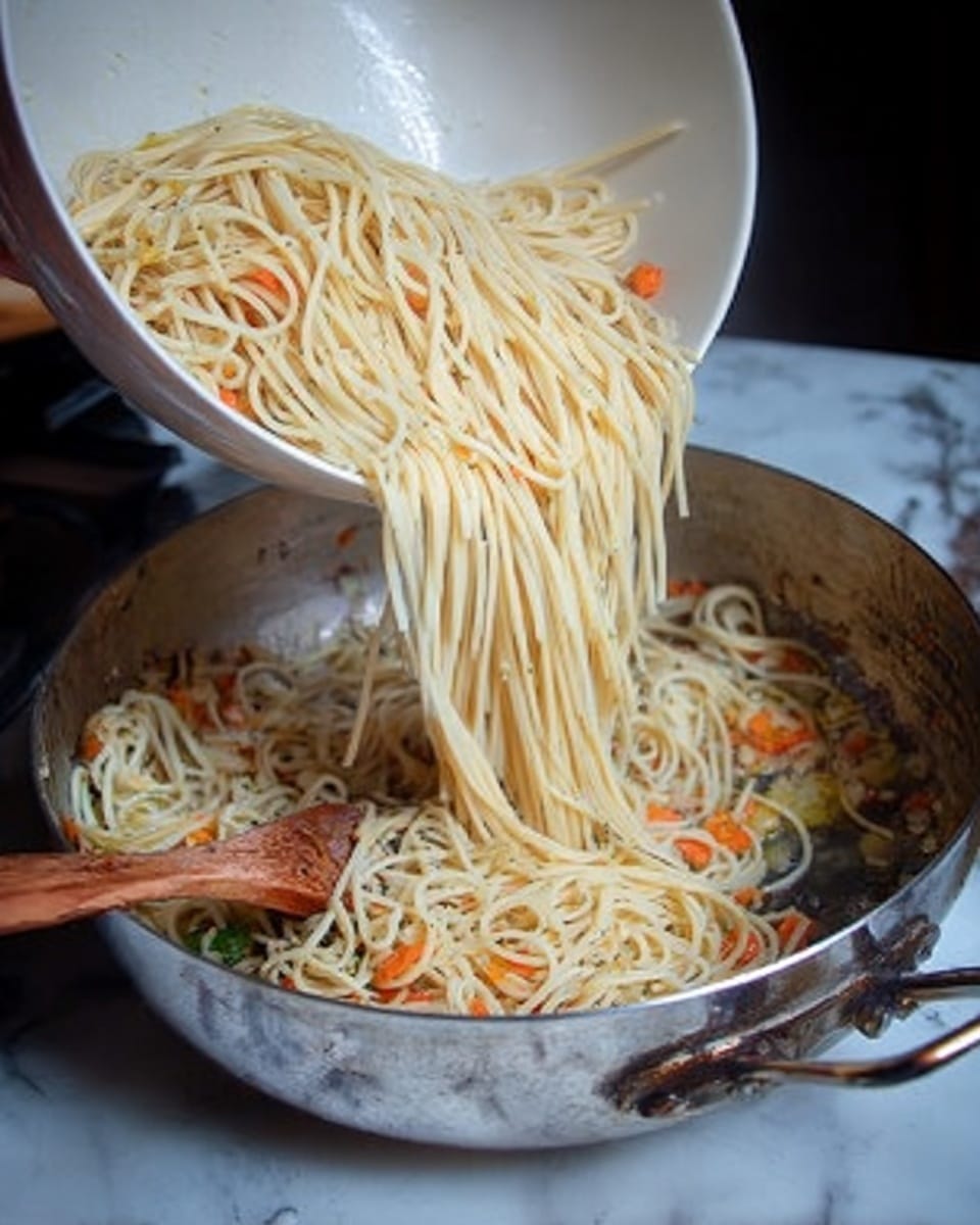 A close-up of a woman's hand pouring cooked spaghetti noodles from a large white bowl into a shiny metal pan with handles, which contains small pieces of orange carrots and bits of green vegetables at the bottom. A wooden spoon rests inside the pan, partially visible under the noodles. The scene is set against a white marbled surface. photo taken with an iphone --ar 4:5 --v 7