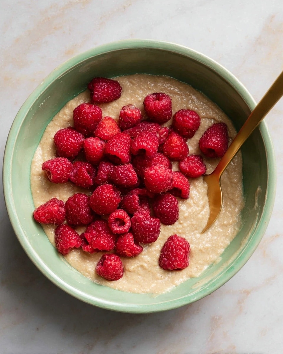 A light green bowl filled with tan-colored thick batter, topped with a pile of fresh, bright red raspberries spread mostly in the center. A gold spoon rests inside the bowl on the right side, partially submerged in the batter. The bowl sits on a white marbled surface showing slight texture. Photo taken with an iphone --ar 4:5 --v 7