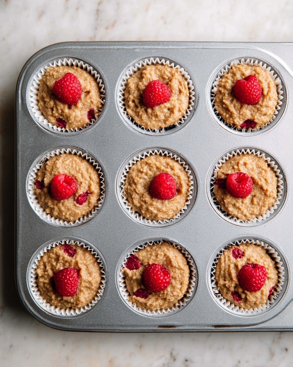 A grey metal muffin tray filled with nine white paper liners, each holding light brown batter with a rough, thick texture, and two bright red raspberries placed on top of each muffin batter. The tray is set on a white marbled surface. The raspberries contrast with the batter, adding a fresh, juicy look. The muffins are evenly spaced in the tray, showing a neat arrangement. Photo taken with an iphone --ar 4:5 --v 7