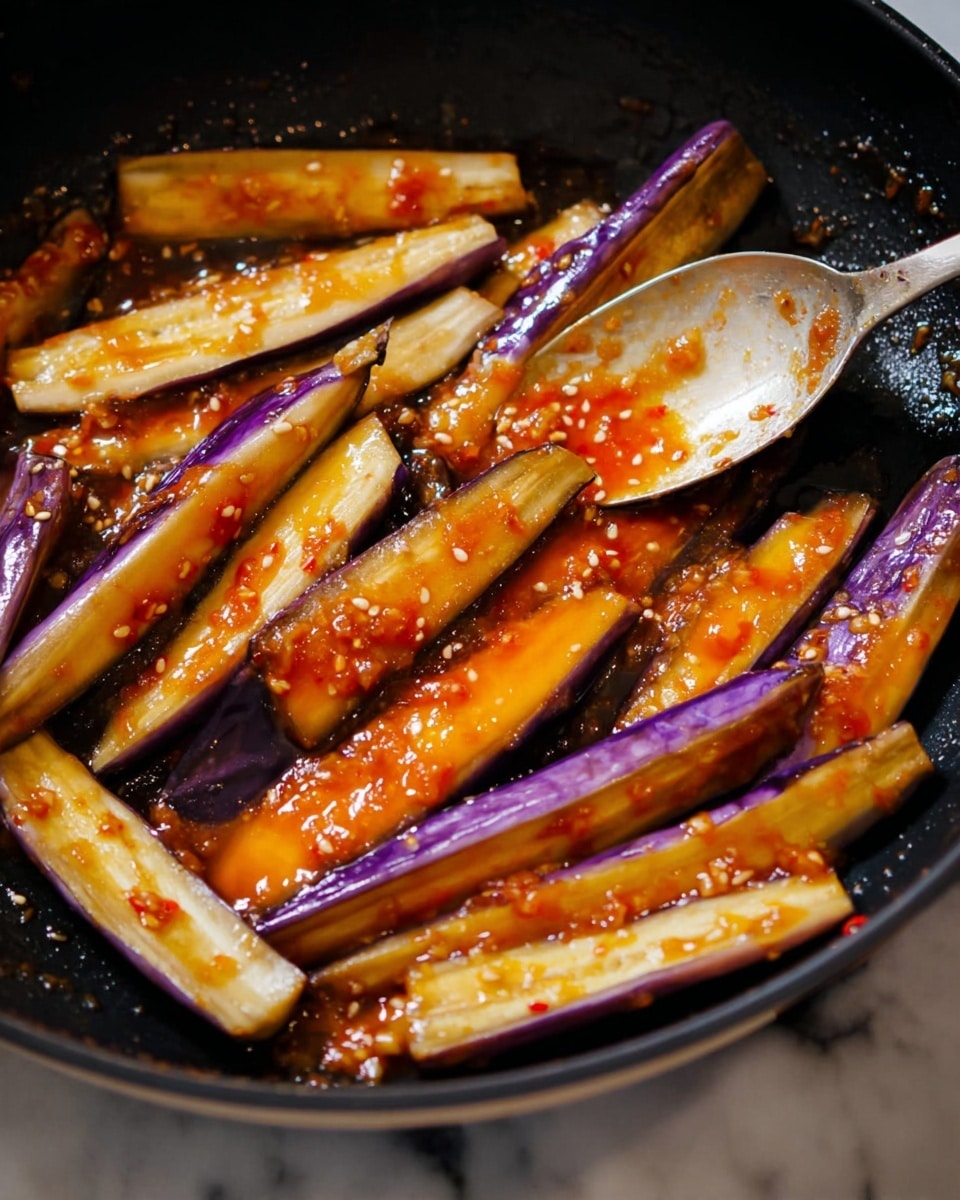 The image shows several long, thin pieces of eggplant cooking in a black pan. The eggplant pieces have a purple skin and light yellow inside. They are covered with a shiny, orange-red sauce with some small seeds and bits, making them look slightly sticky but not thick. In the top right corner, a metal spoon rests in the pan, partly covered with the same sauce. The pan is dark and contrasts with the bright colors of the eggplant and sauce. The background surface is white marble. photo taken with an iphone --ar 4:5 --v 7