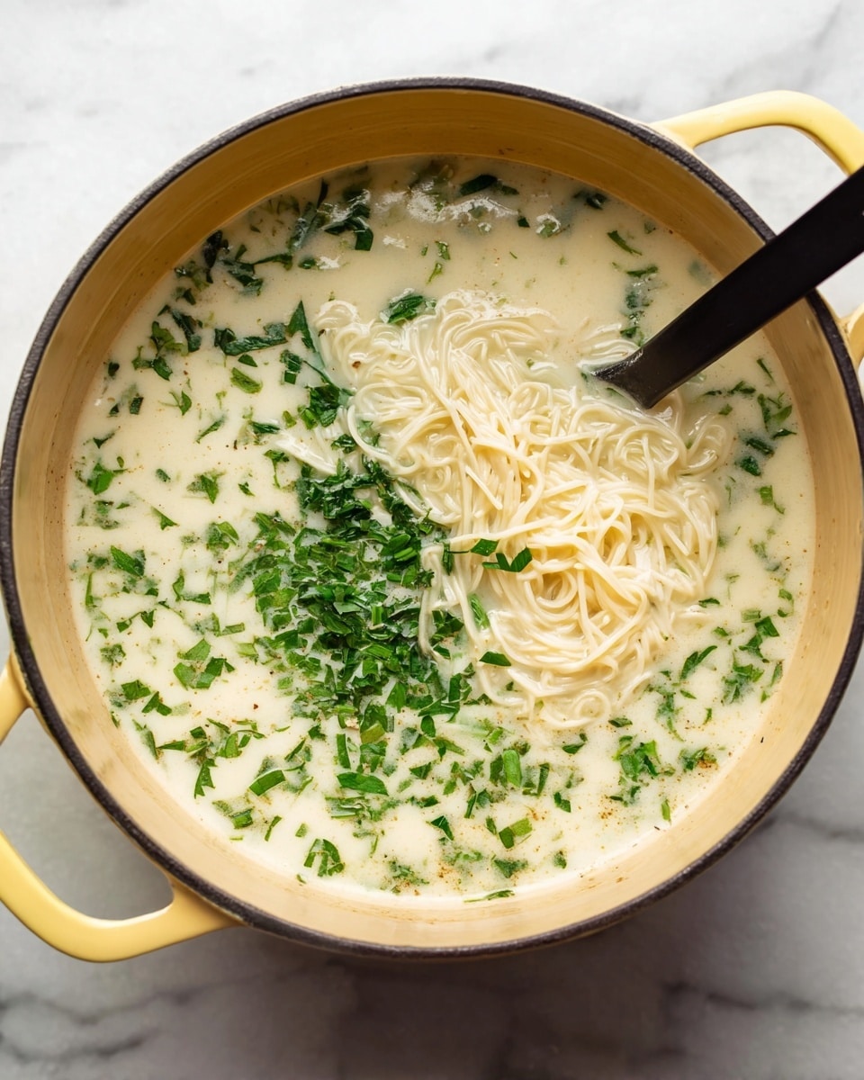 A yellow pot filled with a creamy white soup contains a tangle of thin white noodles lifted by a black spoon on the right side; the soup is dotted with small, chopped bright green herbs evenly spread throughout the liquid. The pot sits on a white marbled surface, and the color contrast between the pale soup, vivid herbs, and black spoon is clear. photo taken with an iphone --ar 4:5 --v 7