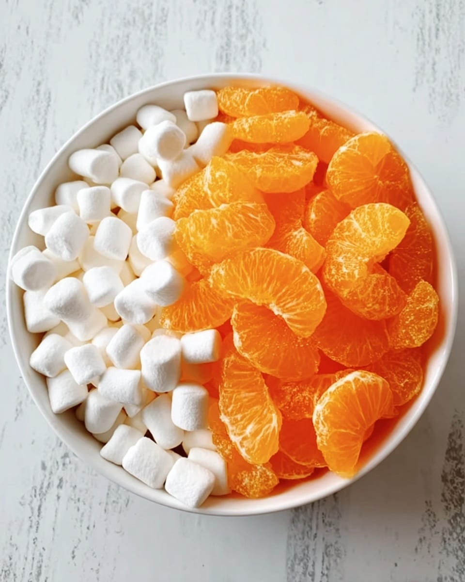 A white bowl filled with two main layers visible from the top. One half of the bowl contains bright orange mandarin orange slices with a juicy and slightly shiny texture. The other half holds small, soft, white mini marshmallows packed closely together, creating a fluffy and smooth layer beside the orange fruit. The bowl is placed on a white marbled surface. photo taken with an iphone --ar 4:5 --v 7