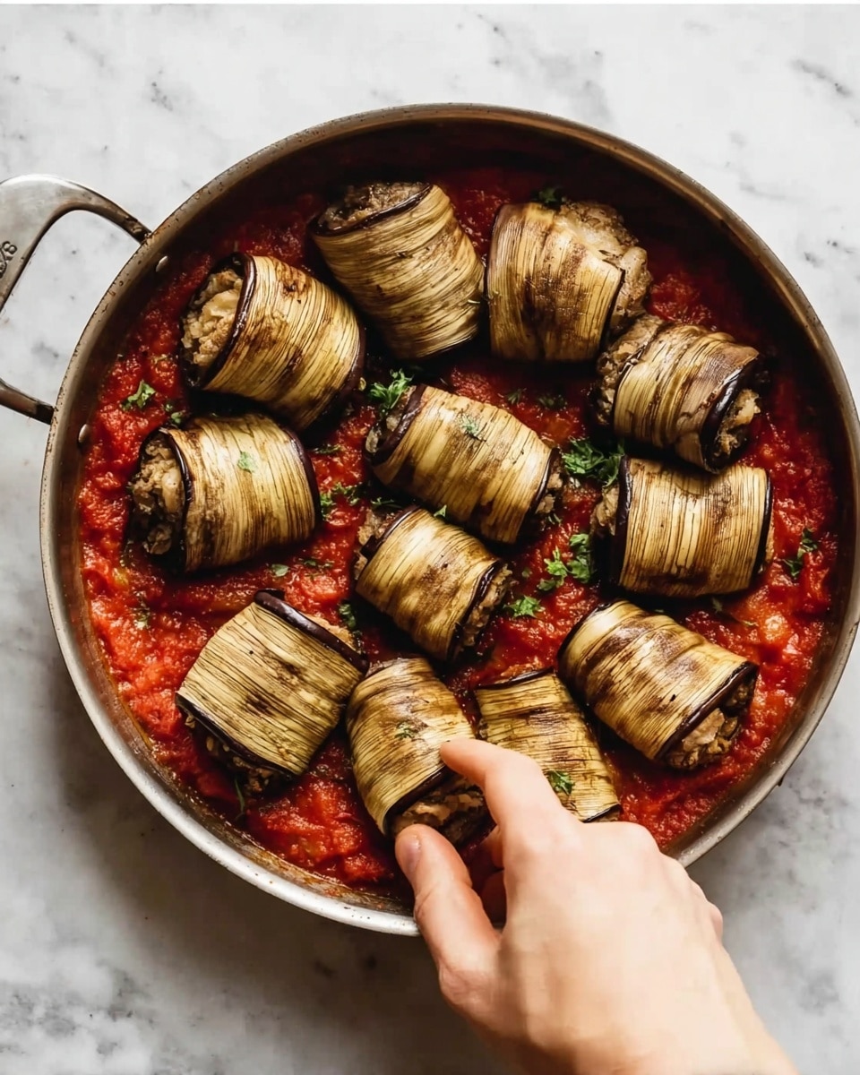 A round metal pan holds a red tomato sauce base with visible chunks of tomato and some green herbs scattered inside. On top of the sauce are ten rolled, browned eggplant slices arranged neatly in a circle, each showing grill marks and a light texture. A woman's hand is seen gently placing one of the eggplant rolls near the edge of the pan. The pan is set on a white marbled texture surface. photo taken with an iphone --ar 4:5 --v 7