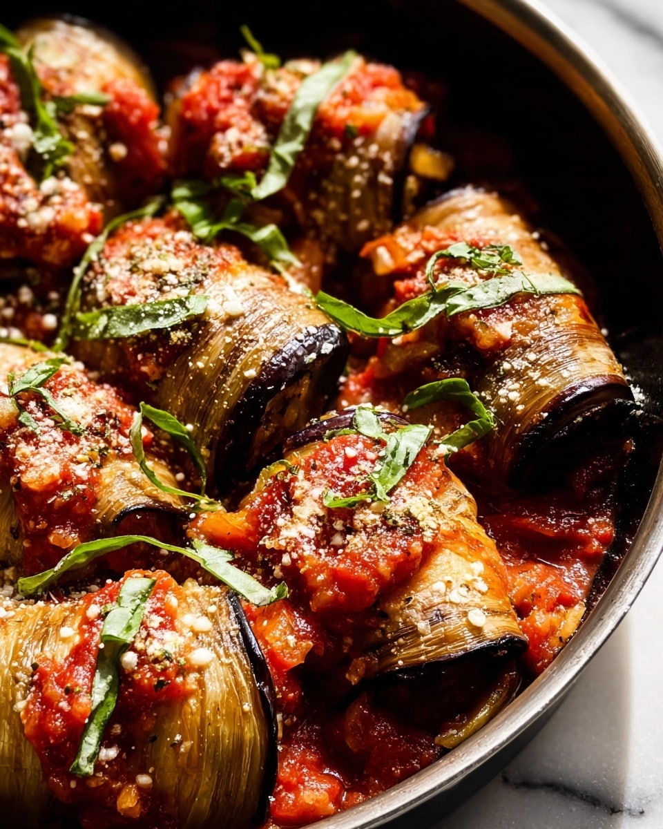 This is a close-up of several rolled eggplant pieces, placed snugly in a dark pan. Each roll is made of thin, grilled eggplant slices with a slightly wrinkled, brownish-purple texture, coated with a chunky red tomato sauce and sprinkled with white grated cheese. Thin strips of fresh green basil lay scattered on top and between the rolls. The pan sits on a white marbled surface with a soft, bright light casting gentle shadows, highlighting the textures and colors. photo taken with an iphone --ar 4:5 --v 7