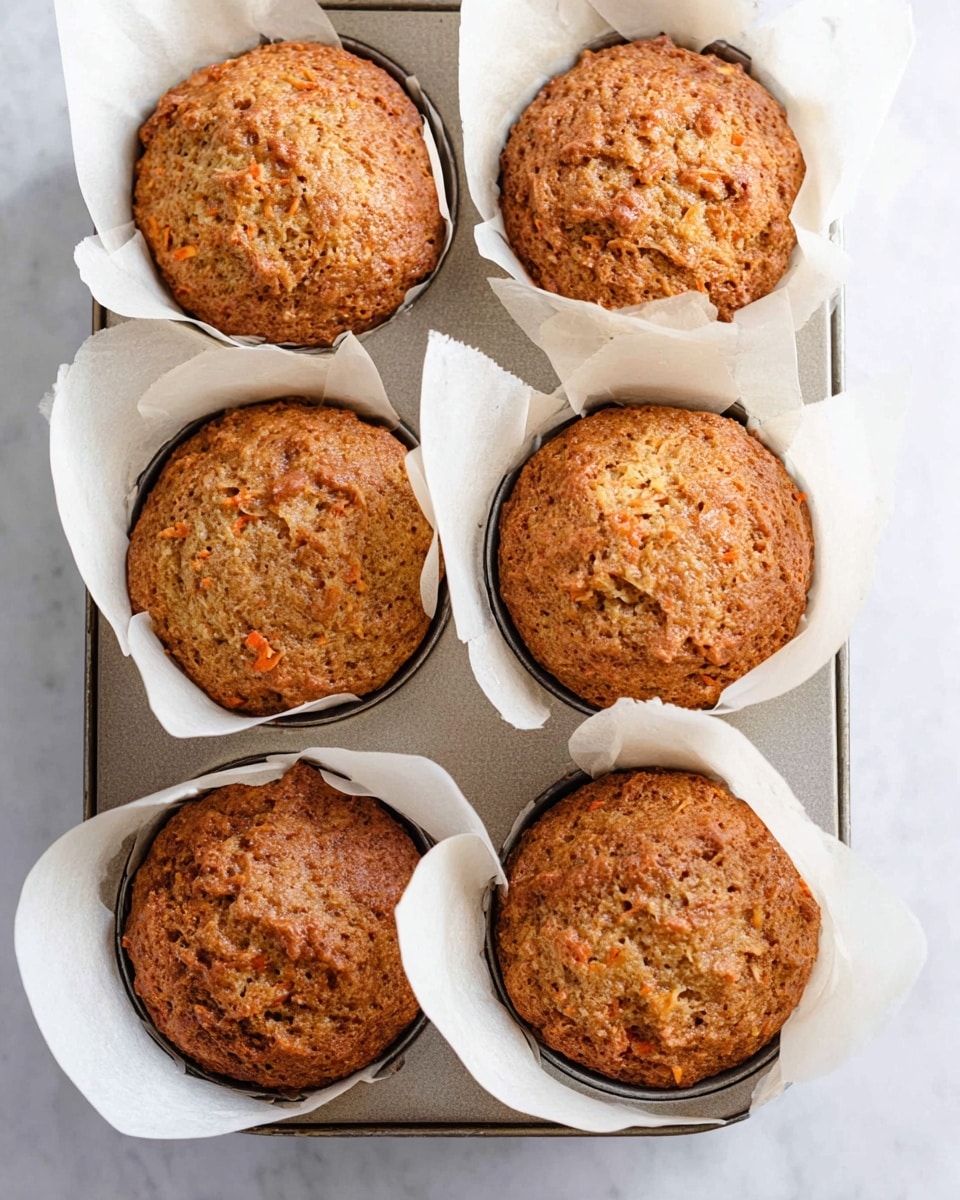 Six brown muffins with a rough and slightly cracked top texture sit in white parchment paper liners inside a metal baking tray. The muffins show small bits of orange, hinting at carrot inside. The tray rests on a white marbled surface. The muffins look freshly baked with a golden brown color and soft crumbly tops. photo taken with an iphone --ar 4:5 --v 7