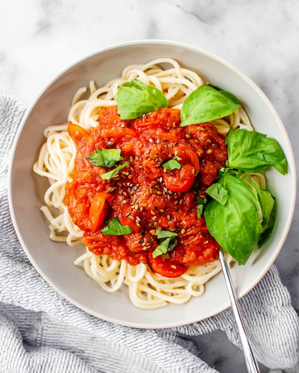 A white bowl holds a dish with two main layers, the bottom layer is light-colored twisted noodles forming a loose nest, while the top layer is a chunky red tomato sauce with visible tomato pieces, scattered herbs, and seeds giving it a textured look; fresh green basil leaves are placed on top, adding color contrast, and a silver fork is partially resting on the noodles on the right side; the bowl sits on a white marbled surface with a soft striped cloth partly visible underneath the bowl. photo taken with an iphone --ar 4:5 --v 7