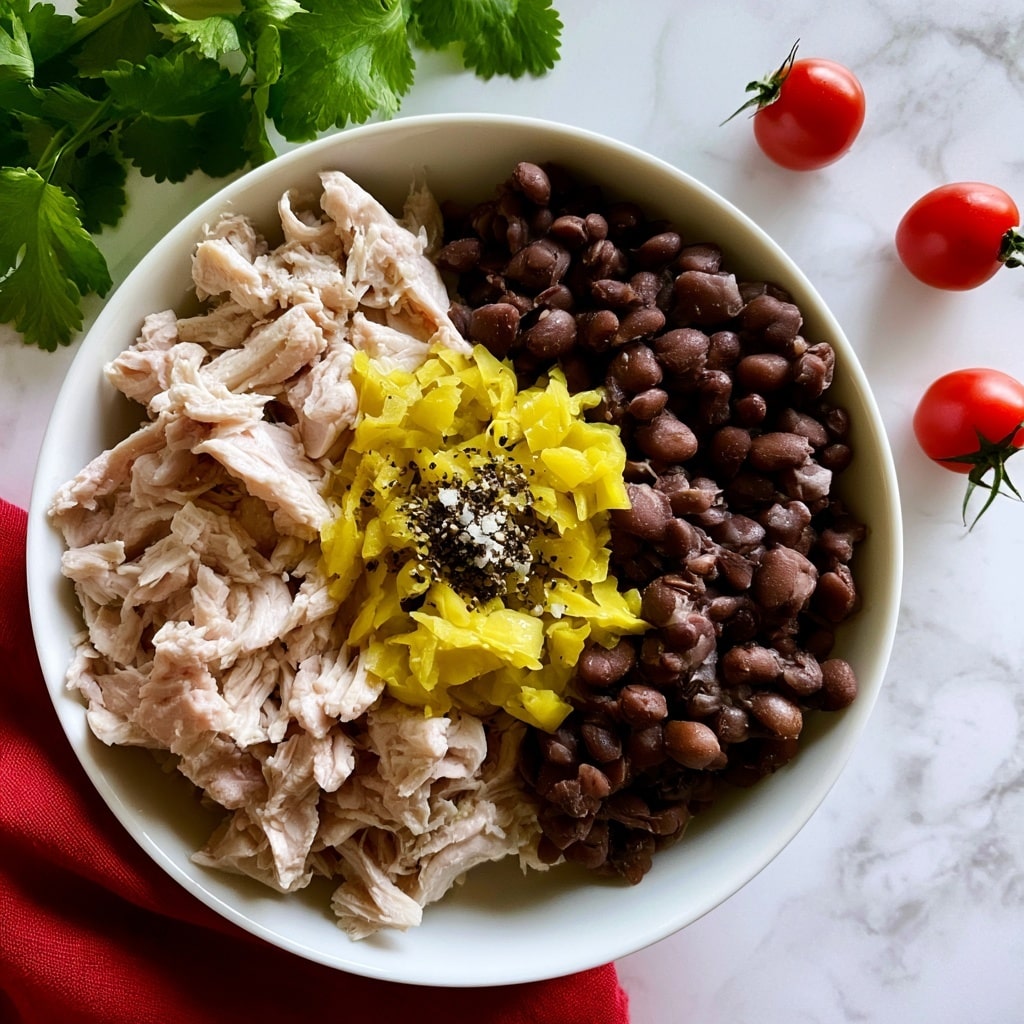 A white bowl on a white marbled surface holds four main layers: on the left side, light pink shredded chicken pieces; on the right, dark brown black beans; in the center, bright yellow chopped pickled peppers; and on top of the middle, a small pile of black pepper and white salt specks. Around the bowl are fresh green cilantro leaves at the top left and three small red cherry tomatoes with green stems on the right side on the white marbled surface. A red cloth is placed under the bowl, peeking from the bottom left corner. photo taken with an iphone --ar 4:5 --v 7