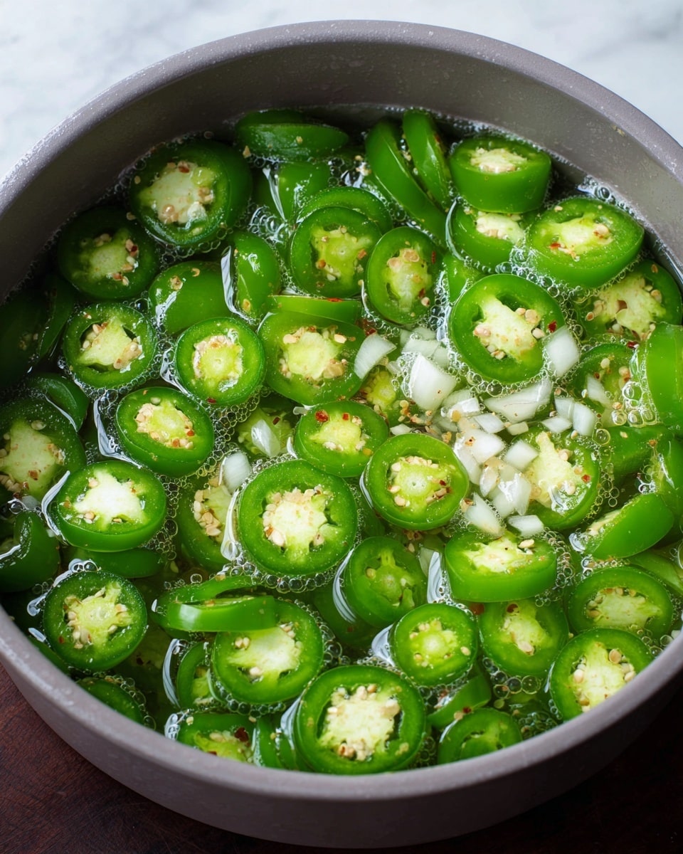 A close up of a light gray pot filled with many bright green jalapeño slices floating in clear bubbly water, mixed with small bits of white onion scattered in the center and throughout. The jalapeños have smooth shiny skins with visible seeds, and tiny and light brown spices sprinkled on top. The pot is set on a white marbled surface. photo taken with an iphone --ar 4:5 --v 7