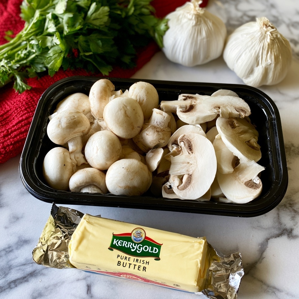 A black plastic container filled with whole and sliced white mushrooms sits on a white marbled surface. To the upper left of the container, a bunch of green leafy parsley rests on a red textured cloth. Near the top right corner, two whole bulbs of garlic with white papery skin are placed side by side. At the bottom part of the image, a stick of wrapped butter labeled