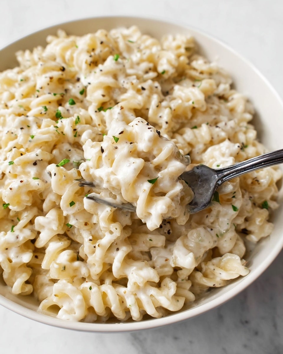This image shows a close-up of creamy pasta in a white bowl. The pasta has a curly shape with a smooth, thick white sauce that coats every piece evenly. Small green herb specks and black pepper flakes are scattered throughout the sauce, adding subtle color and texture. A silver fork is partly dipped into the pasta, lifting some noodles at the center right of the bowl. The background is a white marbled surface, keeping the focus on the creamy pasta dish. Photo taken with an iphone --ar 4:5 --v 7