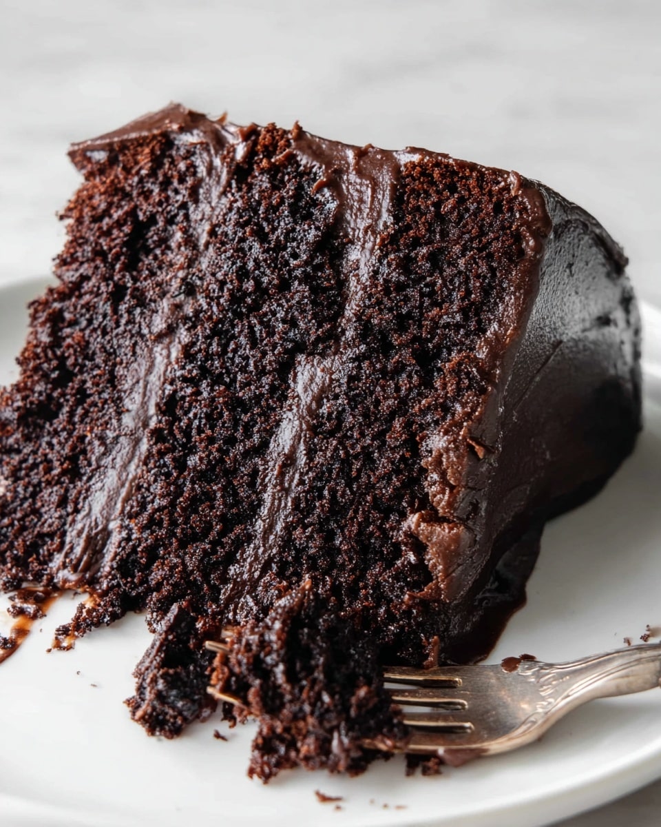 A close-up view of one thick slice of dark chocolate cake on a white plate, showing one single rich chocolate layer with a rough and moist texture, and a shiny smooth dark chocolate frosting covering the top and sides. The focus is on the cake’s soft crumb inside, with a silver fork resting at the plate's edge with some chocolate bits on it. The background is a white marbled texture. photo taken with an iphone --ar 4:5 --v 7
