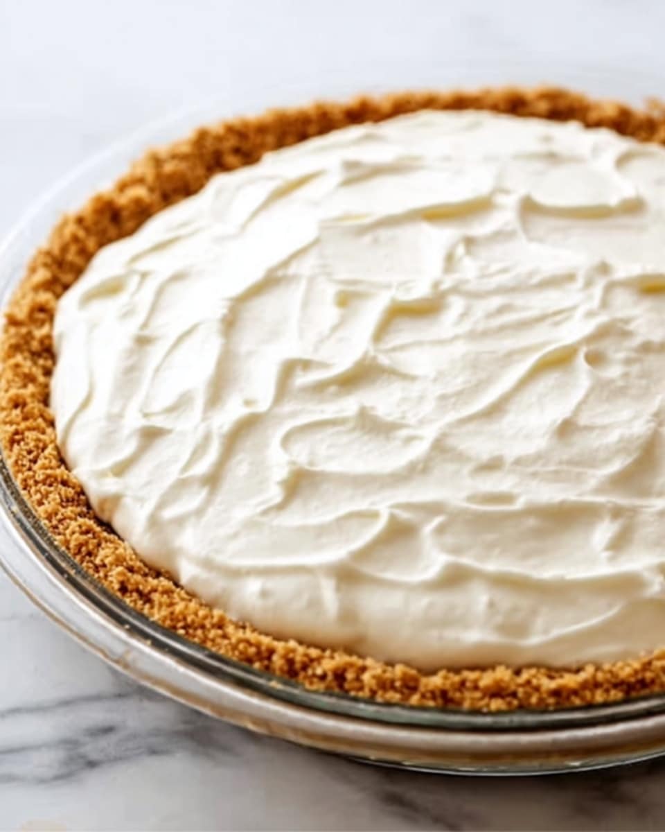 A close-up of a round pie with two layers, sitting on a white marbled surface. The bottom layer is a golden brown crumbly crust that forms a thick edge around the pie. The second layer, filling the entire crust, is a thick, creamy white topping with a smooth and slightly swirled texture. The pie is placed in a clear glass dish. Photo taken with an iphone --ar 4:5 --v 7