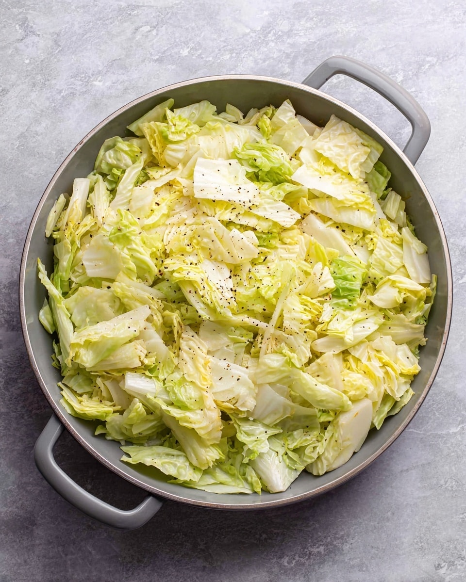 A round gray pan filled with large pieces of light green cabbage layered loosely in no specific order, sprinkled with small black pepper flakes evenly over the top. The cabbage pieces vary in shades from pale yellow-green to a soft bright green, showing a mix of smooth and slightly wrinkled leaf textures. The pan has two handles on each side, and it sits on a white marbled surface. Photo taken with an iphone --ar 4:5 --v 7