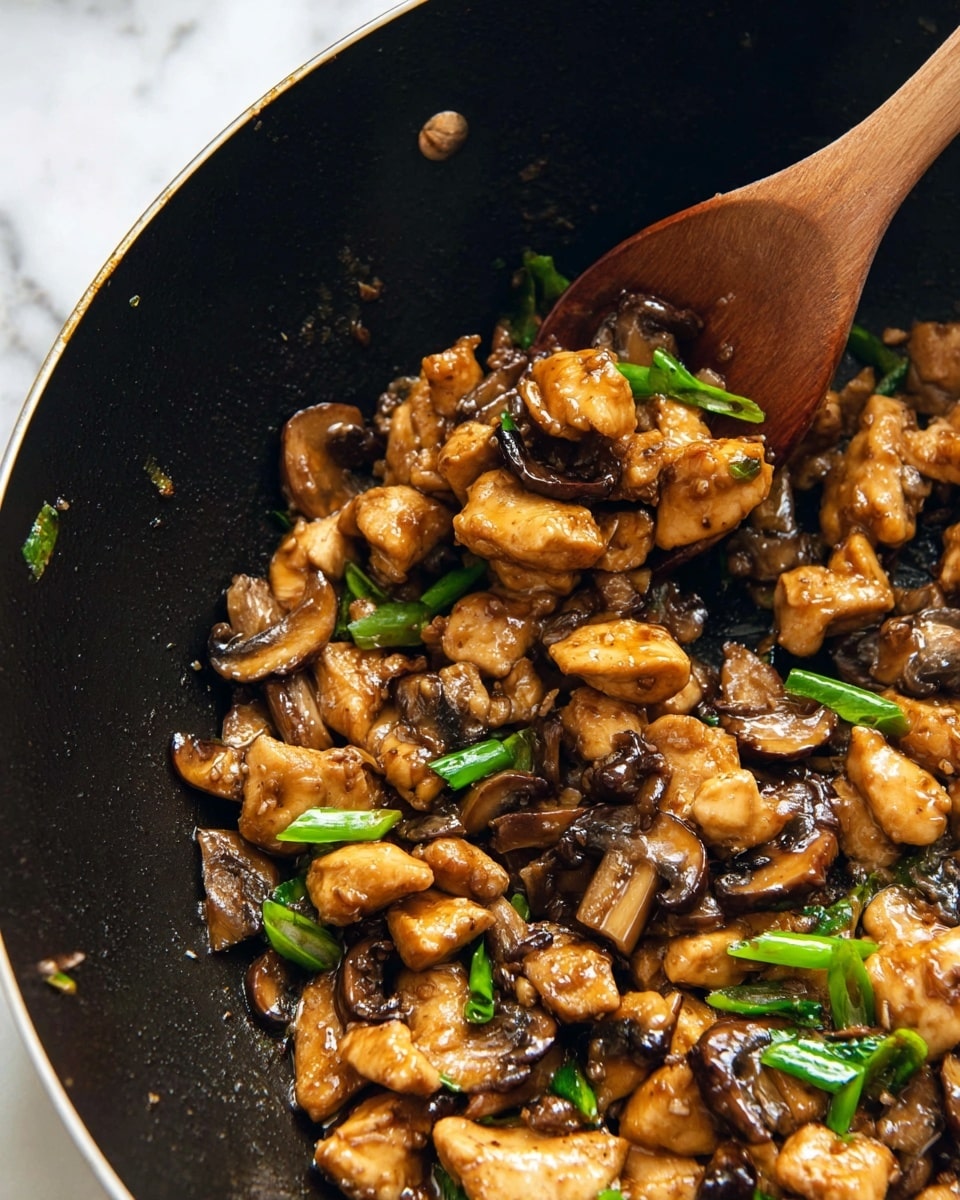 The image shows a close-up of a black wok filled with a stir-fry dish made of small golden brown pieces of chicken, dark brown and light brown sliced mushrooms, and bright green onion slices scattered evenly throughout. The chicken pieces have a slightly shiny, saucy texture that coats them well, blending with the mushrooms and greens. A wooden spoon is partially dipped into the stir-fry, lifting some of the mixture, showing the texture and colors clearly. The background surface is a white marbled texture. Photo taken with an iphone --ar 4:5 --v 7