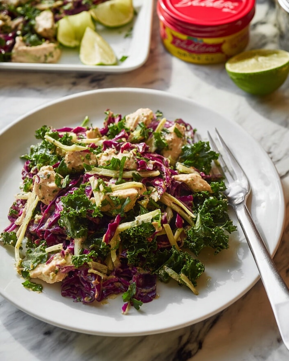 A white plate holds a fresh salad with three main layers: the bottom layer is torn pieces of deep purple cabbage, the middle layer is bright green kale leaves with curly edges, and the top layer features thin pale yellow strips mixed with small chunks of light beige chicken. There are also small sprigs of green herbs scattered throughout the salad, adding extra texture and color. To the right side of the plate, a silver fork rests on the edge. In the background, there is a round red container with a yellow label on a white marbled surface and a square white plate with some salad and lime wedges, all softly out of focus. Photo taken with an iphone --ar 4:5 --v 7