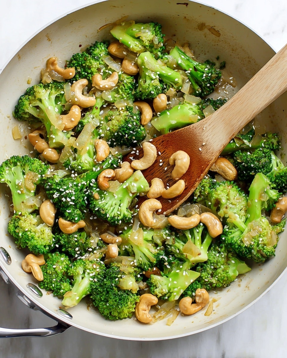 A white frying pan filled with bright green broccoli florets, light golden cashew nuts scattered evenly, and translucent cooked onion slices. The textures are soft but still fresh looking, sprinkled with small white sesame seeds on top. A light wooden spatula rests inside the pan, partially lifting some of the vegetables and nuts. The pan sits on a white marbled surface. photo taken with an iphone --ar 4:5 --v 7