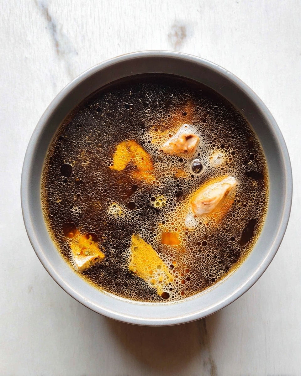 A top view of a light gray bowl filled with clear dark brown broth. The broth has many small oil bubbles on the surface and contains a few irregular pieces of light orange meat submerged in it. The bowl is placed on a white marbled surface. photo taken with an iphone --ar 4:5 --v 7