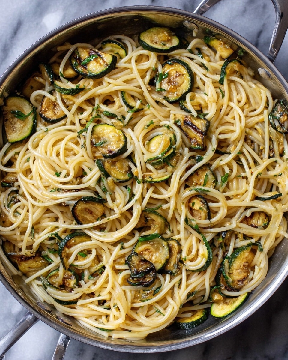 A large silver pan filled with cooked spaghetti noodles mixed with thin slices of grilled zucchini. The spaghetti is light yellow and soft looking, intertwined with dark green and golden brown zucchini slices, some with slight charring. Small bits of green herbs are scattered throughout, adding color contrast. The pan rests on a white marbled surface. photo taken with an iphone --ar 4:5 --v 7