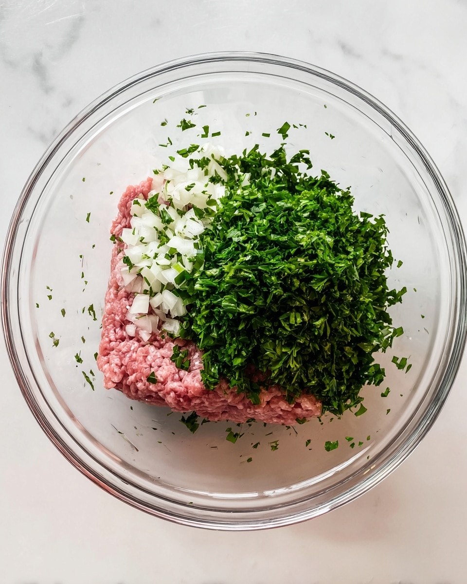 Inside a clear glass bowl on a white marbled surface, there are three main layers of ingredients. The bottom layer is a block of raw ground meat that is pink with a soft texture. On the left side of the meat, there is a layer of small white onion pieces mixed with chopped green herbs. Covering most of the meat on the right and middle is a thick layer of finely chopped bright green parsley. The herbs and onions scatter a bit around the edges of the bowl. The photo was taken with an iphone --ar 4:5 --v 7