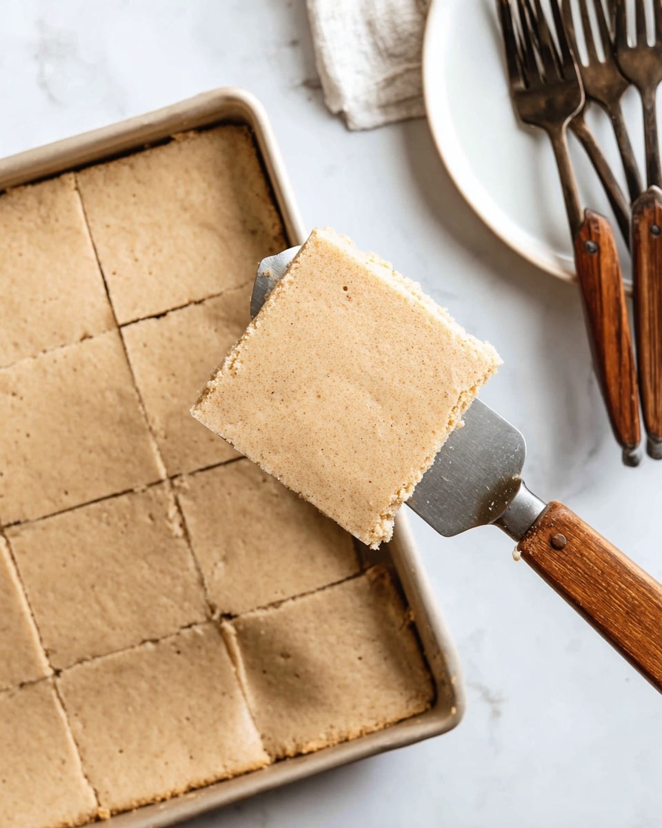 This image shows a single square piece of light brown, thick, soft cake-like dessert with small specks all over, held by a spatula with a wooden handle. The piece is lifted up from a large rectangular baking tray filled with more of the same cake, cut into squares. The edge of a clean white plate sits nearby on a white marbled surface, alongside several folded forks with brown and black handles. The overall look is warm and rustic, with a simple, fresh presentation photo taken with an iphone --ar 4:5 --v 7